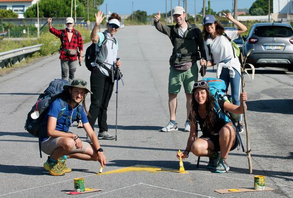 seis peregrinos, alguns a acenar, dois agachados a pintar a estrada