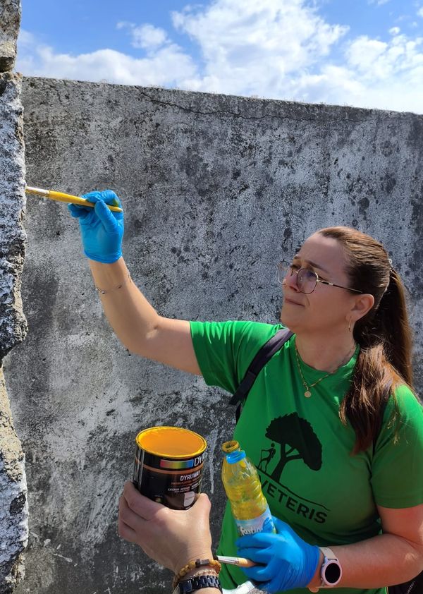 Uma pessoa com um pincel e uma lata de tinta amarela a pintar um muro
